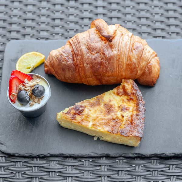 A croissant, quiche slice, and yogurt parfait with berries and granola on a dark slate.