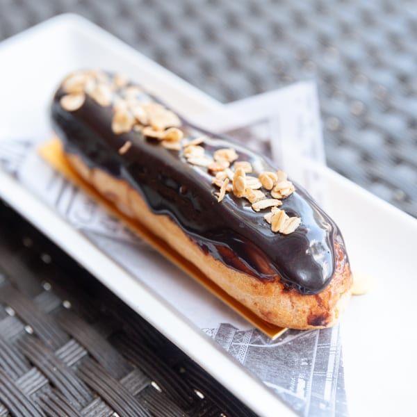 A chocolate eclair topped with nuts on a white plate with a patterned paper liner.