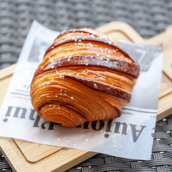 A golden-brown pain au chocolat, dusted with powdered sugar, rests on a newspaper and wooden board.