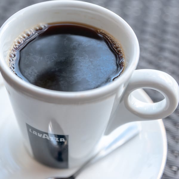 A white Lavazza coffee cup filled with dark coffee and a spoon on a saucer.