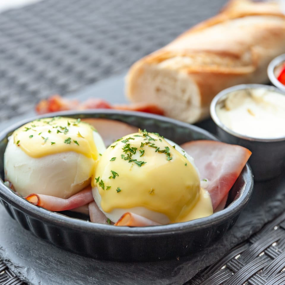 Two eggs Benedict with hollandaise sauce and parsley, served with ham, baguette, and sauces.