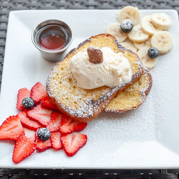 French toast with ice cream, powdered sugar, sliced strawberries, bananas, and blueberries on a white plate.