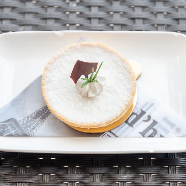 A white tart with whipped cream, a chocolate shard, and rosemary on a French newspaper-patterned napkin.