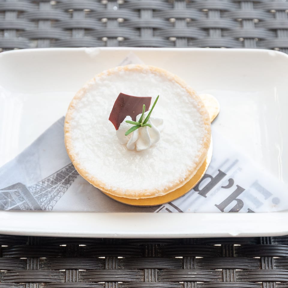 A white tart with whipped cream, a chocolate shard, and rosemary on a French newspaper-patterned napkin.