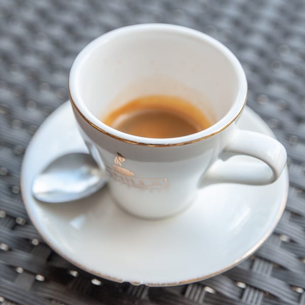 A white espresso cup with gold trim and the Vanilla Espresso logo, on a saucer with a spoon.
